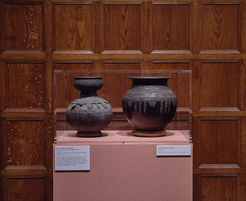 Unknown, Dowry, water or palm wine vessel (1900–1970). Stoneware; Unknown, Pot (1900–1970). Stoneware. Exhibition view: Body Vessel Clay: Black Women, Ceramics & Contemporary Art, Two Temple Place, London (29 January–24 April 2022). © Two Temple Place. Courtesy Two Temple Place. Photo: Amit Lennon Two dark clay pots set in the same exhibition room against another wooden wall. On a pink plinth