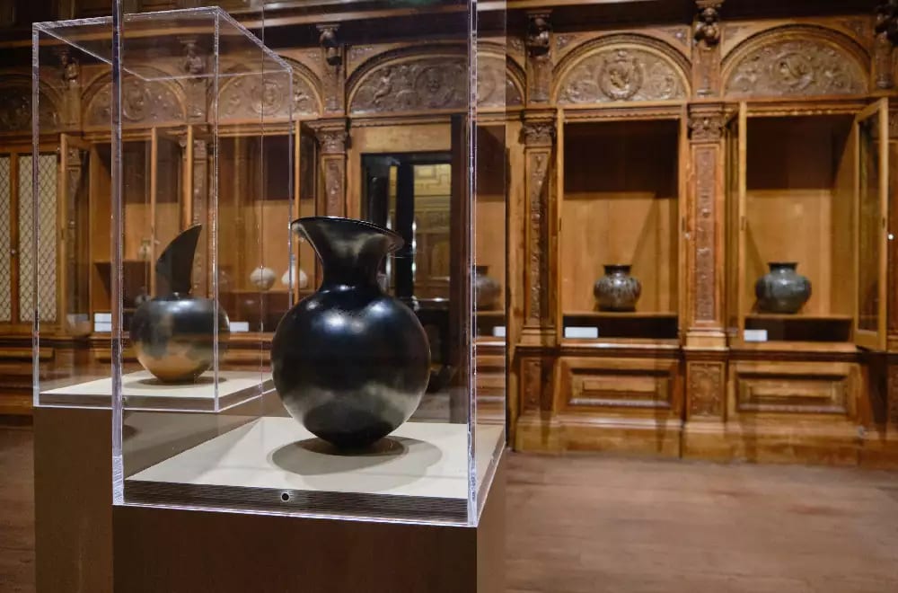 Magdalene Odundo, Symmetrical black pot with angled top (1983). Exhibition view: Body Vessel Clay: Black Women, Ceramics & Contemporary Art, Two Temple Place, London (29 January–24 April 2022). © Two Temple Place. Courtesy Two Temple Place. Photo: Amit Lennon. Black shiny clay pot in glass display cabinet