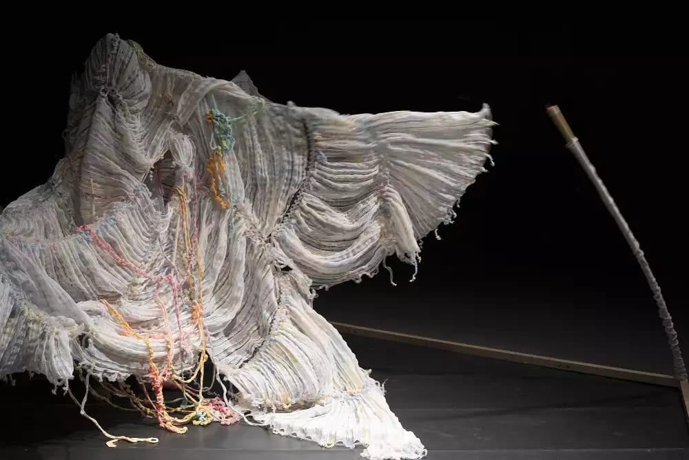 The woman continues to perform, completely beneath the yarn, stretching her arms out wide