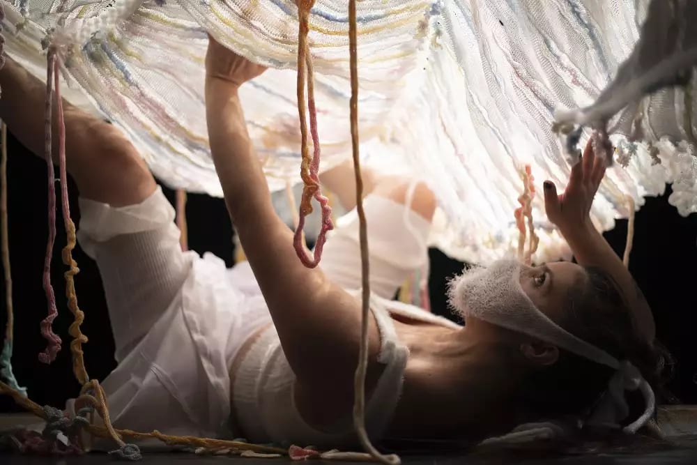 The woman is now lying underneath the yarn, propping it up with her hands and legs