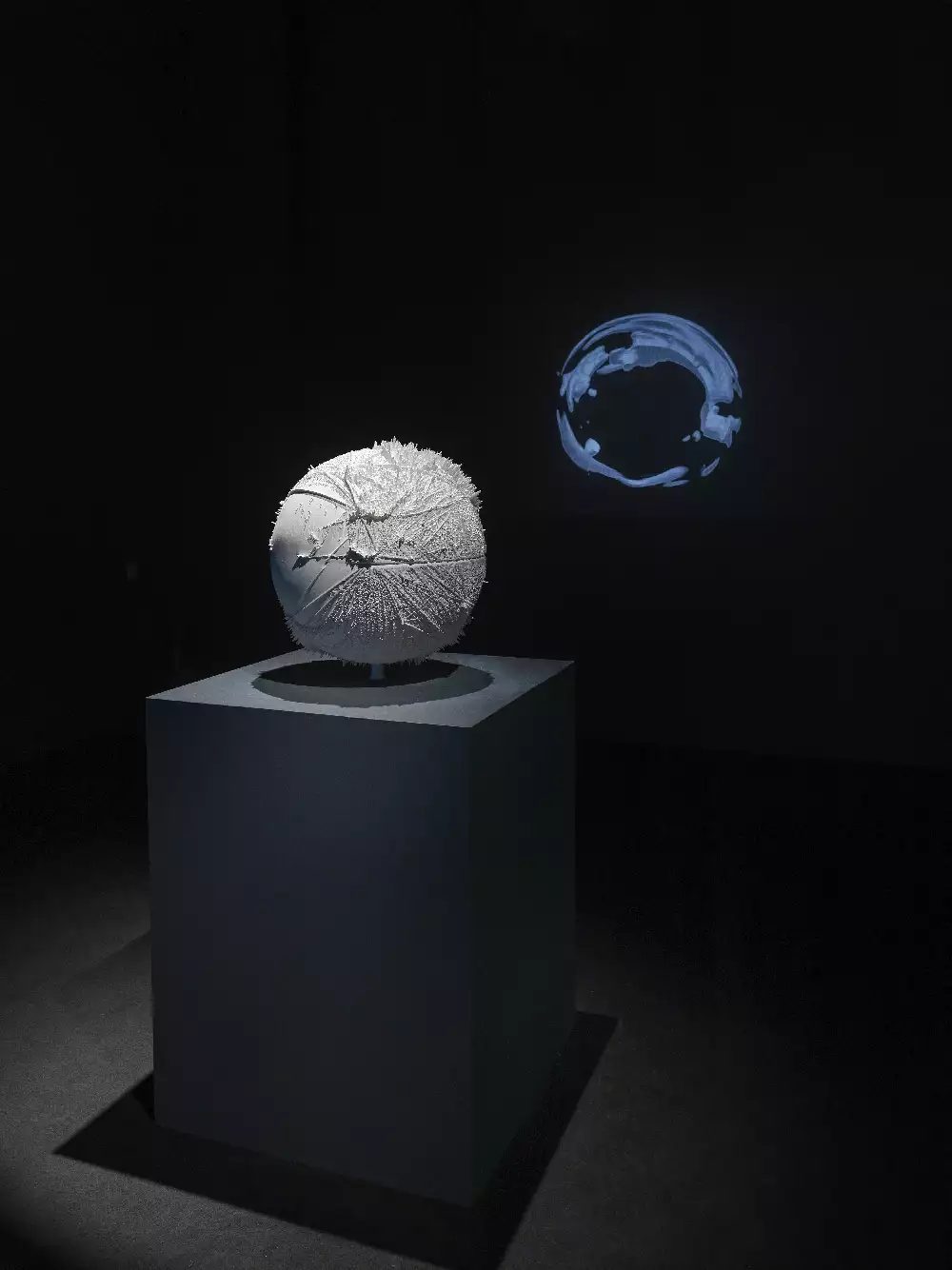 Carbon Topologies by Manifest Data Lab, installed at Weather Engines exhibition, Onassis Stegi, Athens 2022. Image of a grey plinth with a white circular sculpture in a dark room