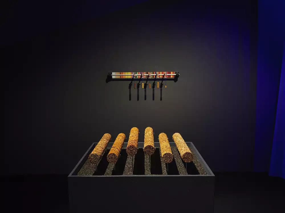 Proxy Climates, by Anca Benera & Arnold Estefan, installed at Weather Engines exhibition, Onassis Stegi, Athens 2022. six orange tubes on a plinth in a dark room with a spotlight shining onto them